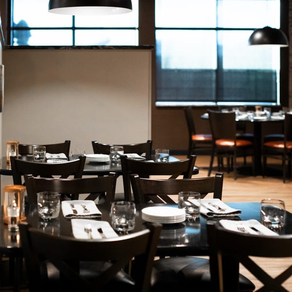 Set tables with dark chairs, white plates, and cutlery in Ovest Via's dining room.