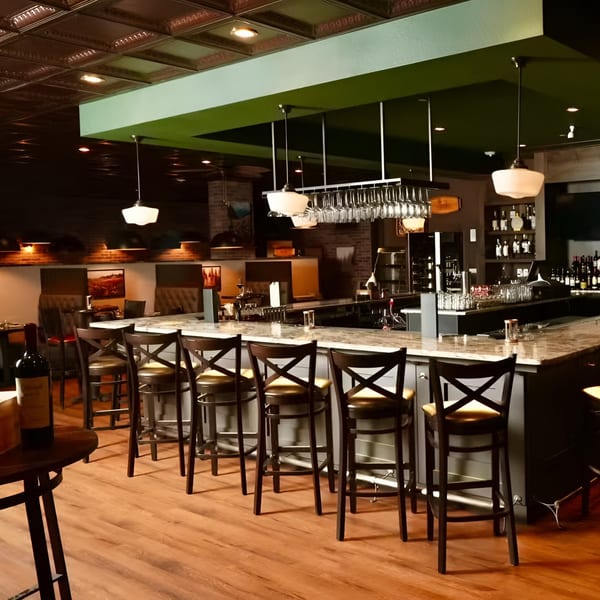Ovest Via's elegant bar area features a long counter with stools, hanging wine glasses, and warm lighting.