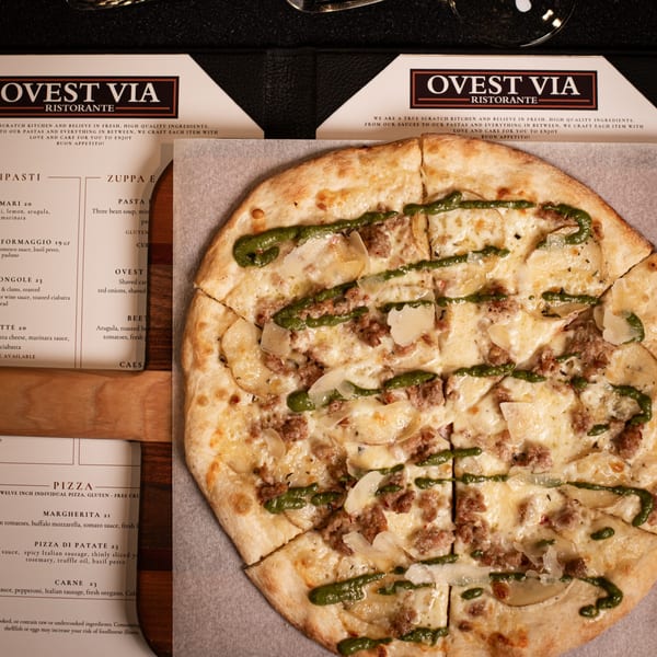 Ovest Via's Pizza di Patate with sausage, potatoes, and pesto drizzle next to a menu.