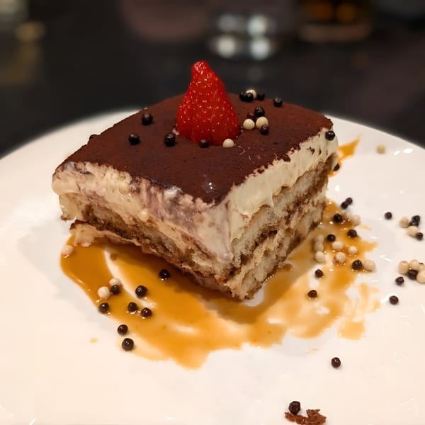 A slice of Ovest Via's tiramisu, topped with cocoa powder, a strawberry, and chocolate pearls.
