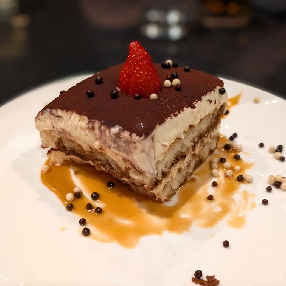 A slice of Ovest Via's tiramisu, topped with cocoa powder, a strawberry, and chocolate pearls.