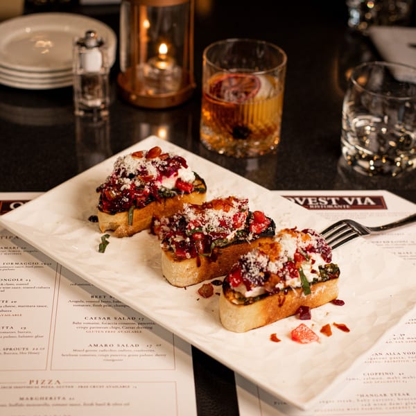 Three bruschetta appetizers with red toppings and cheese, a drink, and menu on a dark table.