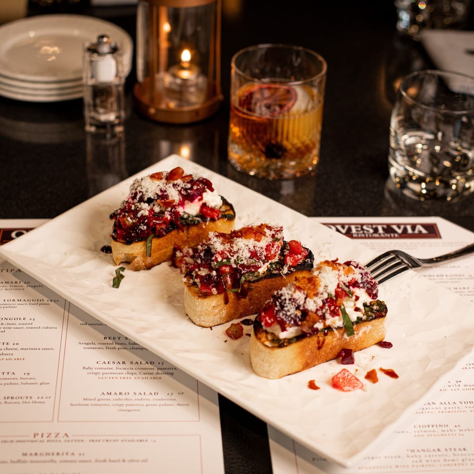 Three bruschetta appetizers with red toppings and cheese, a drink, and menu on a dark table.