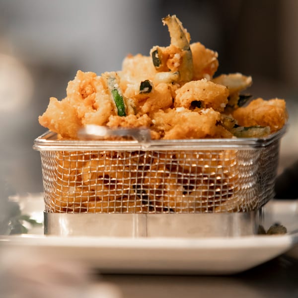 Crispy fried calamari and vegetables in a metal basket, served on a white plate.